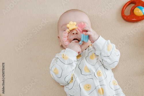 Baby lying on bed and gripping teething ring with both hands. Child chewing toy for teething relief. Exploring textures while soothing sore gums. Sensory discovery at home.