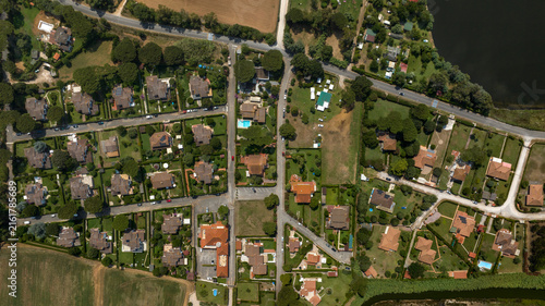 Aerial top down view of a quiet residential neighborhood with detached houses, green gardens, private swimming pools and tree lined streets bordering a dark lake, showing suburban layout from above.