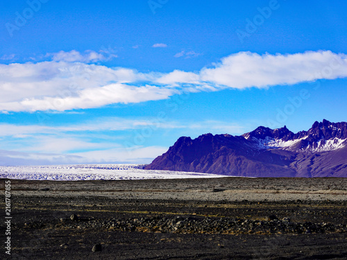 Fotografía A vast glacier tongue and rugged mountain ridges rise beyond a flat black sand p