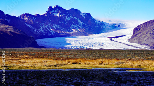 Cuadro en lienzo A sweeping glacier flows between rugged, snow-dusted mountains beyond a golden g