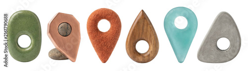 Collection of diverse location map pins made from various natural and industrial materials like stone, copper, felt, wood, glass, and concrete, arranged in a horizontal line