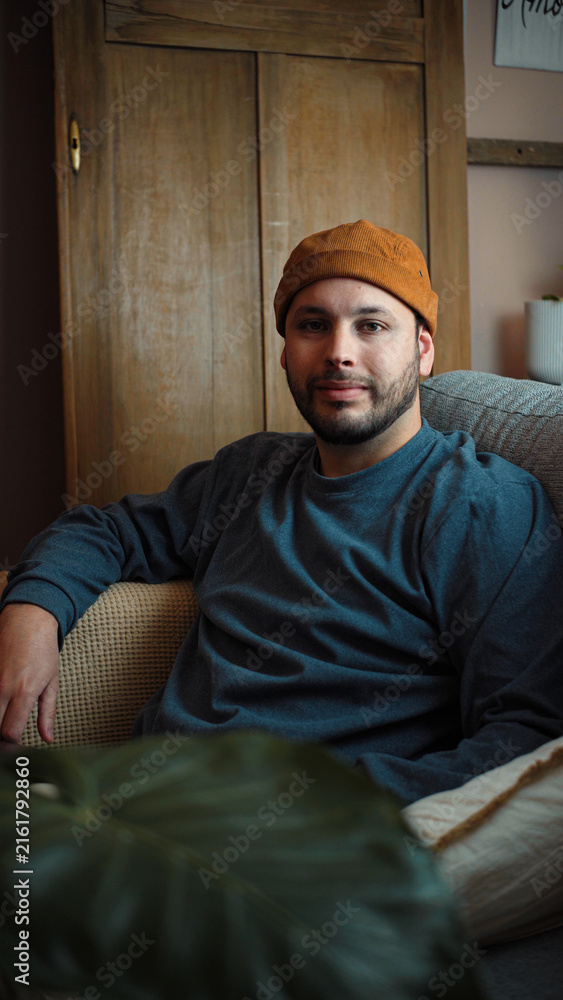 Fototapeta premium Young Latino man sitting on his sofa and looking directly at the camera, creating a natural and approachable portrait in a comfortable home setting.