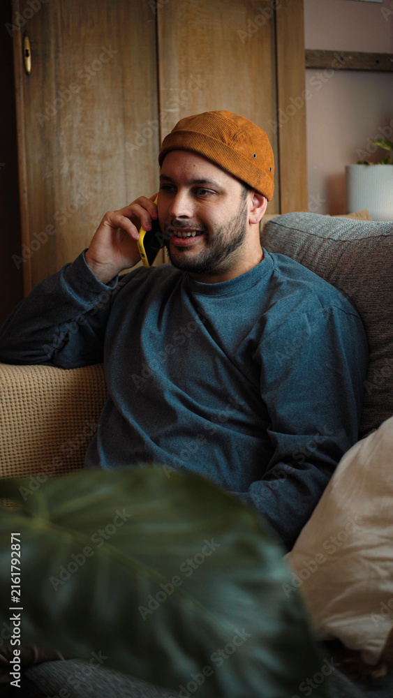 Fototapeta premium Happy young Hispanic man sitting on his sofa and talking on the phone, smiling during a cheerful conversation in a comfortable home setting.
