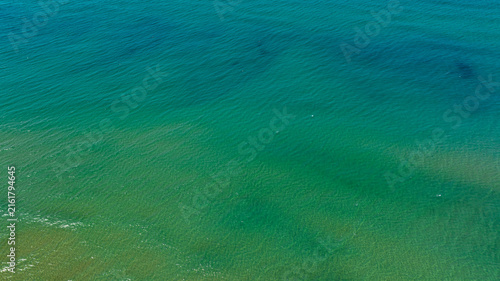 Aerial close up view of crystal clear turquoise and emerald green sea water with gentle ripples and shallow sandy seabed visible, creating abstract natural texture pattern in a sunny morning.