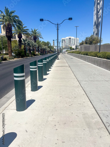 Row of green safety bollards along an empty concrete sidewalk with palm trees and city buildings under a clear blue sky