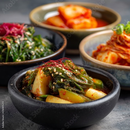 A collection of various traditional fermented vegetable side dishes served in small ceramic bowls, featuring spicy seasonings, fresh herbs, and sesame seeds on a textured dark background.