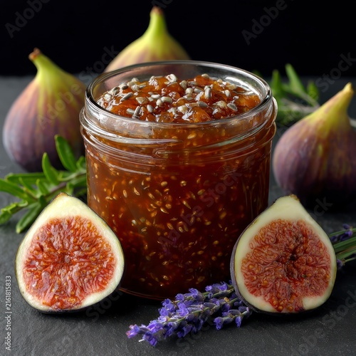 A glass jar filled with fresh fig jam, surrounded by whole and halved ripe purple figs, lavender sprigs, and green herbs on a dark slate surface.