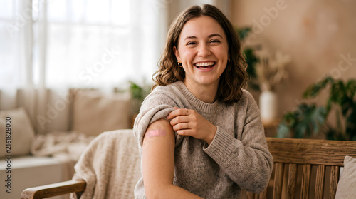Smiling young woman showing adhesive bandage on her upper arm after getting vaccinated or receiving a shot at a healthcare clinic office.