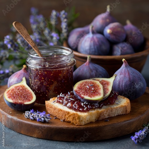 A slice of toasted bread topped with fig jam and lavender flowers, served on a wooden board with a glass jar of jam and whole fresh figs in the background.