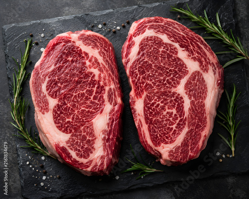 Canvastavla Two raw marbled ribeye steaks resting on a black stone board with fresh green ro