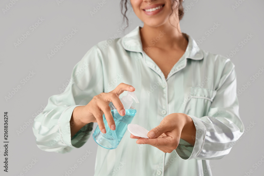 Fototapeta premium Young African-American woman applying micellar water on cotton pad against grey background, closeup
