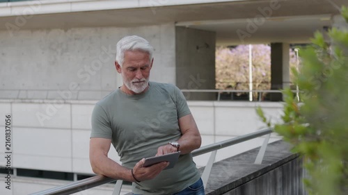 Smiling senior man relaxing outdoors with a digital tablet near a modern residential building. Active retirement and technology concept.