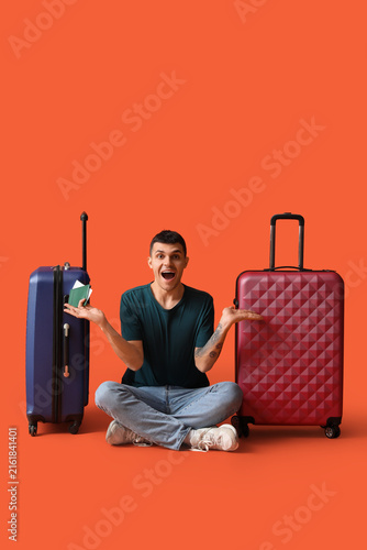Surprised male tourist with suitcases and passport sitting on orange background