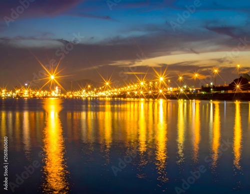 City lights sparkle across a body of water at dusk, reflecting in the serene surface