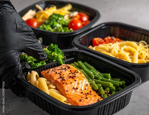 Close-up of gloved hand holding a prepared food container with salmon, pasta, and vegetables