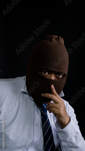 Obraz na plátně An enigmatic Asian male wearing a crisp white shirt and a blue striped tie conceals facial features using a brown knitted balaclava against a dark background