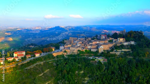 Mysterious aerial pedestal down shot from Santa Vittoria in Matenano with roads, buildings, trees, thickets, churches, belltower, clocktower, walls, surrounded by ravines and sky with various clouds