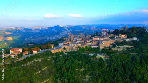 Verdant aerial push-in shot from Santa Vittoria in Matenano, the high-density town centre, a vintage human settlement surrounded by trees, thickets, ravines, typical Marche hillscapes, cloudy skies
