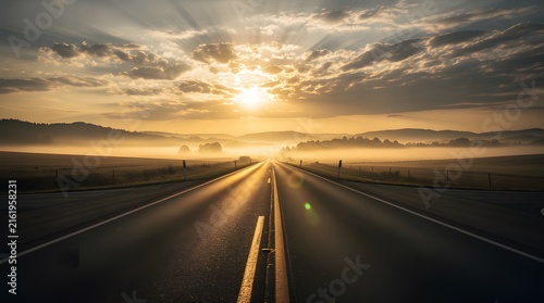 An empty asphalt road stretching towards the horizon during a foggy sunrise with dramatic clouds.