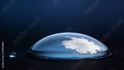 Artistic macro of water droplet reflecting sky and clouds on dark background, with tiny secondary droplet beside it. Great for creative and abstract concepts.