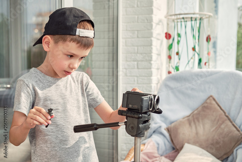 A focused young boy in a gray t‑shirt and black cap sets up a camera on a tripod indoors, holding a toy figure. Soft daylight streams through the window, illuminating the cozy room 