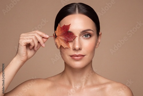 Beautiful woman holding an autumn maple leaf over her eye, natural makeup and glowing skin