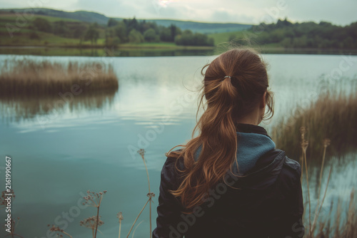 Rear view of a thoughtful young woman standing by a serene lake, contemplating nature in a tranquil landscape
