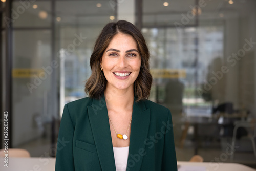 Head shot of young Mexican businesswoman dressed in elegant jacket, smiling, looking at camera standing alone in contemporary meeting room, posing for photo. Office portrait of female CEO or executive
