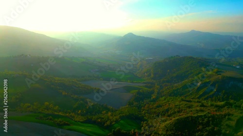 Mystical aerial pullback truck right pan right shot near Santa Vittoria in Matenano, hazy misty Sibillini Mountains in the distance, imbued with spiritual sunlight, ravines and hills in the foreground
