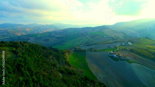Soothing aerial pedestal up push-in truck right pan left shot near Santa Vittoria in Matenano, tiny roads stretching atop lofty hillscapes, cars passing by, hazy sunlight imbuing the view 