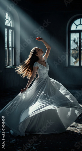 Young woman in white dress spinning gracefully in dark abandoned room illuminated by dramatic sunbeams