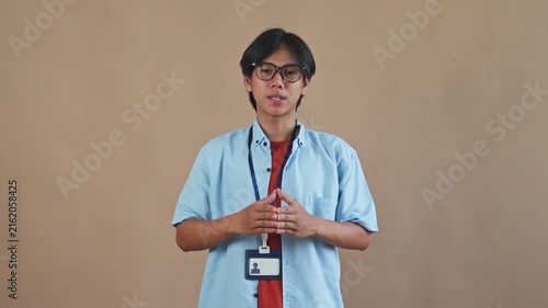 Young Asian male wearing glasses and ID card pers speaking and gesturing directly to camera in studio setting