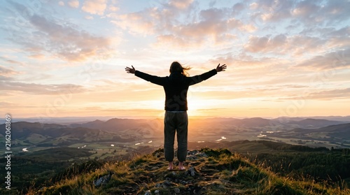 Person standing on mountain peak with arms raised at sunrise, enjoying scenic valley view, freedom and achievement lifestyle concept in nature landscape