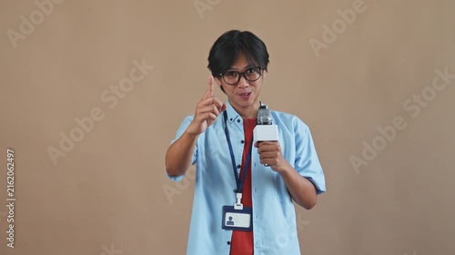Young Asian male reporter with glasses holding microphone while gesturing and talking cheerfully in studio setting