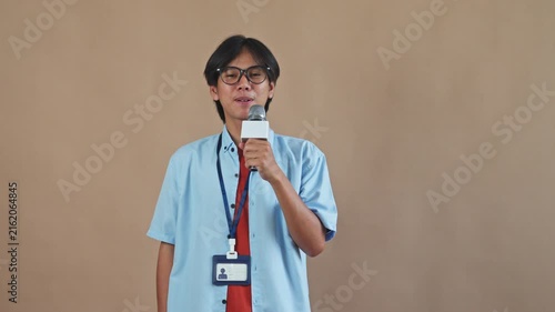 Young Asian male reporter with glasses holding microphone and talking cheerfully to camera in studio setting