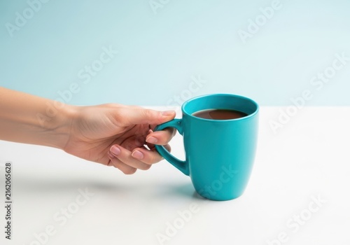 A hand holding a bright turquoise ceramic mug filled with dark coffee against a light blue background