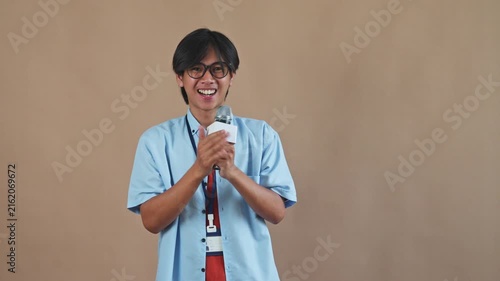 Young Asian male reporter with glasses holding microphone while laughing and speaking happily in studio setting