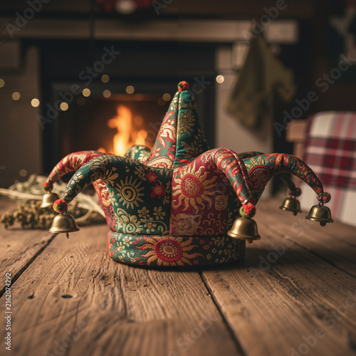 Colorful jester hat with bells on wooden table in cozy interior by the fireplace