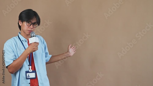 Asian male reporter holding microphone and gesturing toward copy space while speaking in studio setting
