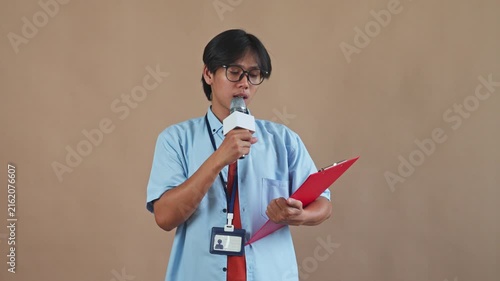Young Asian male reporter with glasses holding microphone and checking red clipboard while speaking in studio