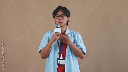 Young Asian male reporter holding microphone while placing his hand over his chest and panting nervously in studio