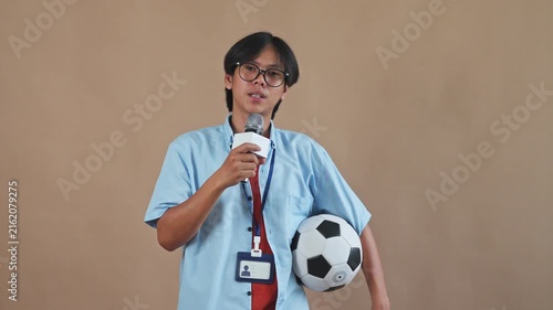 Asian sports reporter with glasses holding microphone and soccer ball while speaking in studio setting