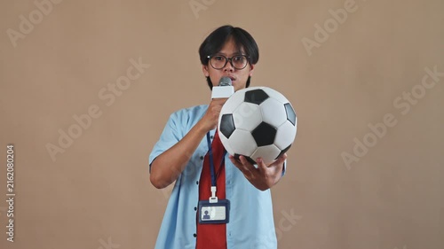 Asian sports reporter holding microphone and showcasing soccer ball while speaking excitedly in studio