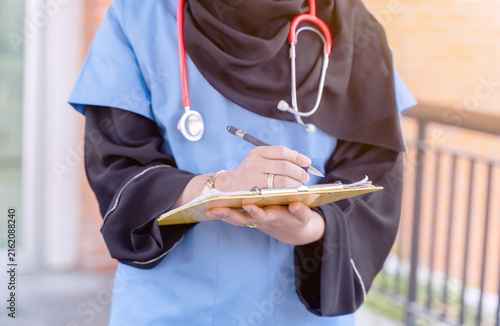 Closeup and crop female Muslim doctor in Abaya hijab traditional dress works at modern clinic office to checking of work list on clipboard