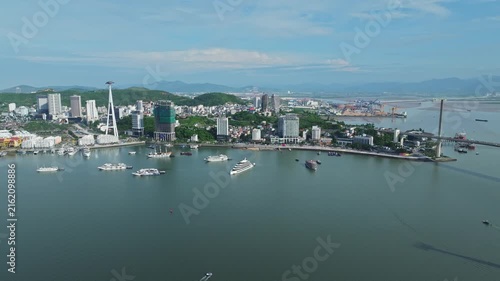 Drone footage overlooking Ha Long city, harbor, boats, waterfront buildings, mountainous coastline, and bridge in Quang Ninh, Vietnam.