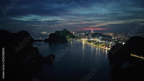 Drone footage of Ha Long Bay at night, featuring dark limestone islands, illuminated waterfront, calm water reflections, and a glowing Quang Ninh cityscape.