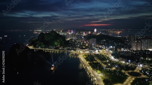Drone view of illuminated Ha Long City along the waterfront at night, featuring urban lights, a scenic bay, coastal roads, parks, and distant skyline.