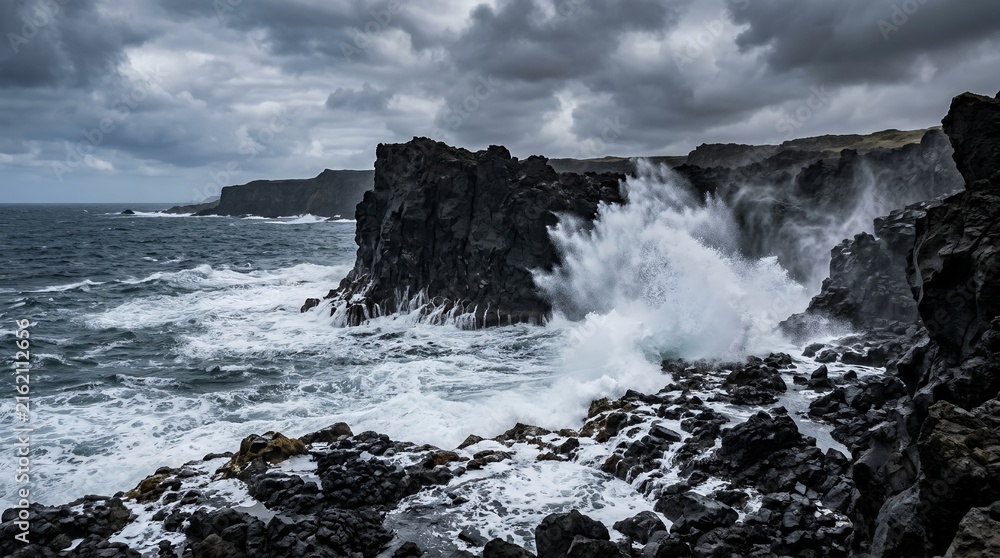 Fototapeta premium Dramatic ocean waves crashing against dark volcanic rocks under a moody stormy sky on a rugged coastal landscape.