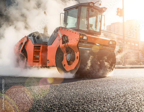 Heavy orange road roller compacting asphalt on a sunny construction site, creating steam and dust, symbolizing urban development, infrastructure projects, and heavy machinery at work.