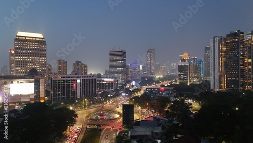 Aerial drone view of Jakarta's Sudirman business district at night featuring illuminated skyscrapers, glowing office buildings, vibrant city lights, and heavy traffic after office hours.